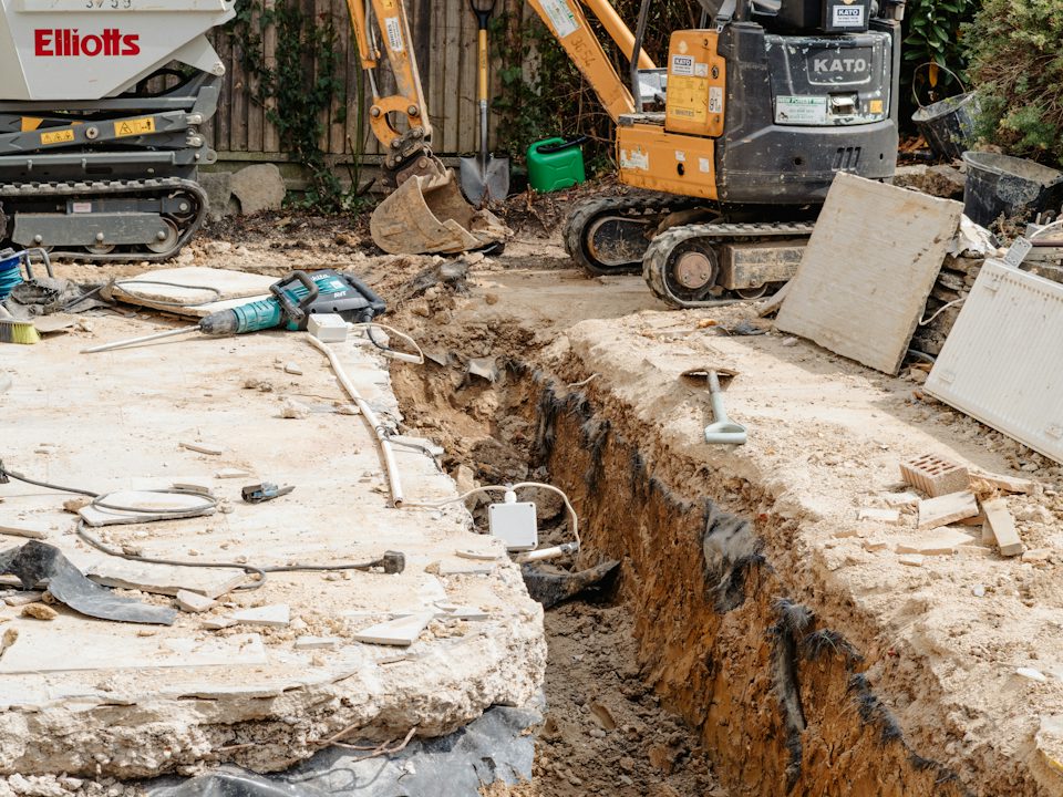 A trench dug in a backyard, with a mini excavator behind it